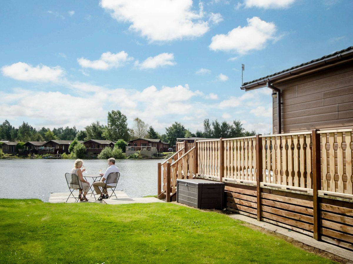 A couple sitting at a small garden table on a paved area of lawn outside a timber lodge, with the lake and more lodges on the far bank behind.