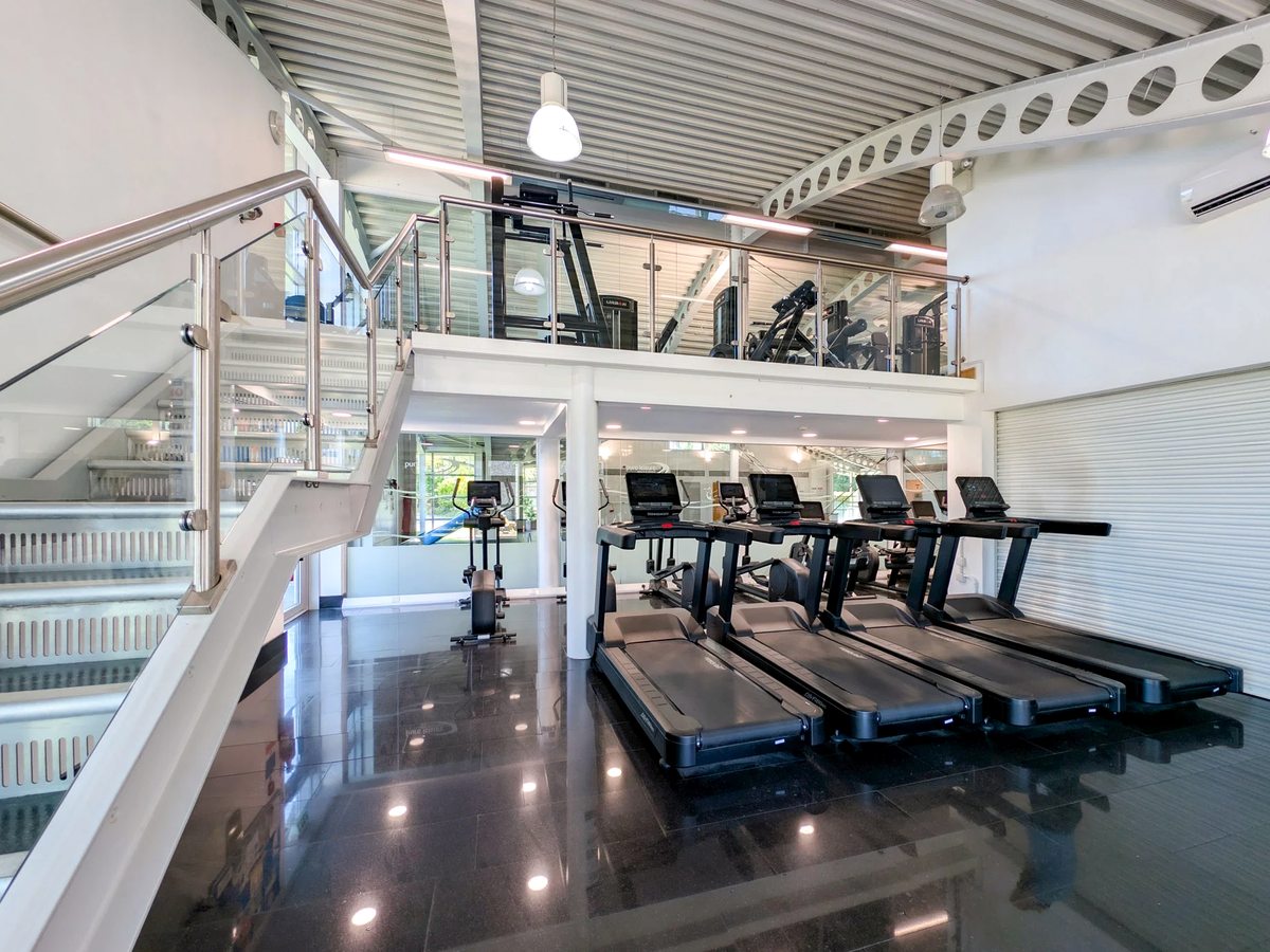 A gym with a row of treadmills on the ground floor and resistance machines on a glass-railed mezzanine above, reached by a white staircase.