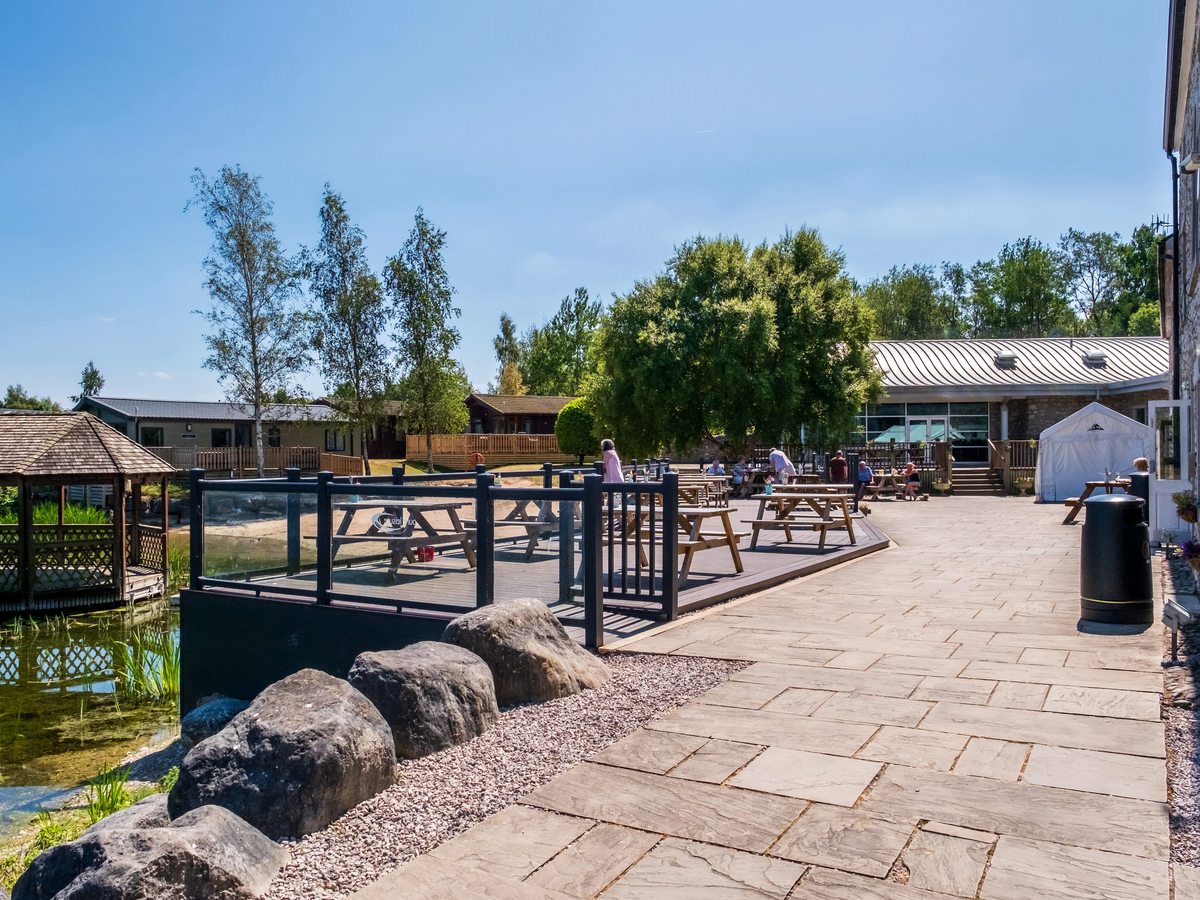 A paved courtyard beside a reedy pond, with a railed timber deck of picnic tables, a wooden gazebo over the water, a low glass-fronted leisure building on the right and lodges among trees beyond.