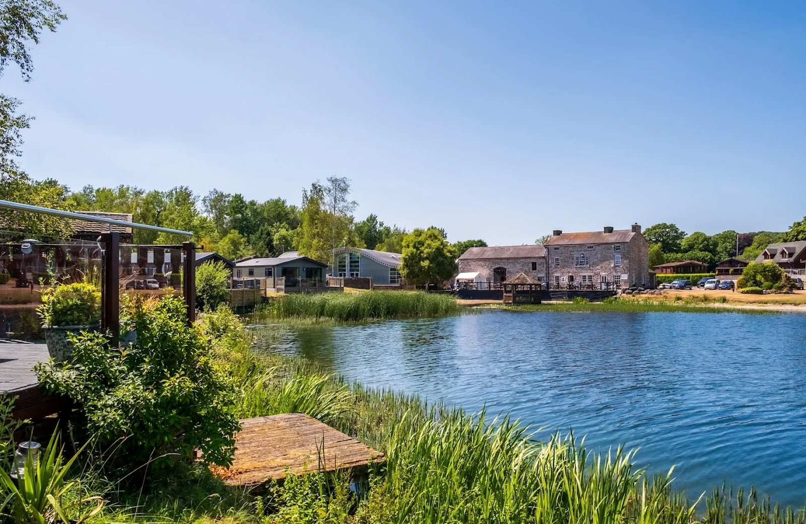 A wide private lake with reeds along its edge, a stone bar and restaurant building and a curved-roofed leisure centre on the far bank, and timber lodges among trees to the right.