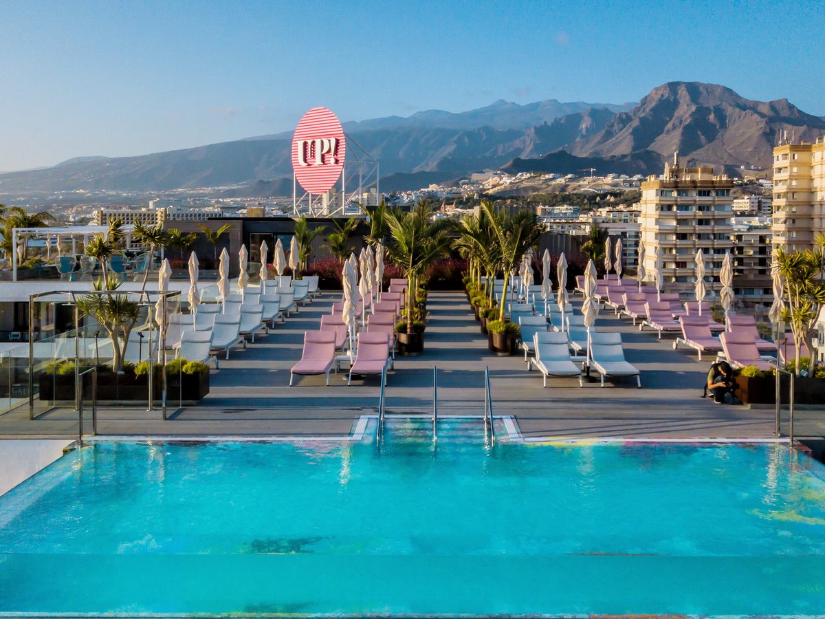 A long rooftop infinity pool with rows of pink and white sun loungers, potted palms, a pink UP! sign and mountains beyond.