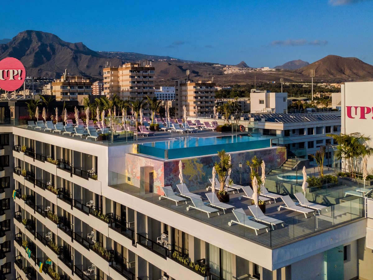 The upper floors of a white high-rise hotel with planted balconies, and on the roof a glass-sided infinity pool, rows of pink and white sun loungers and a pink UP! sign, with dry mountains beyond.