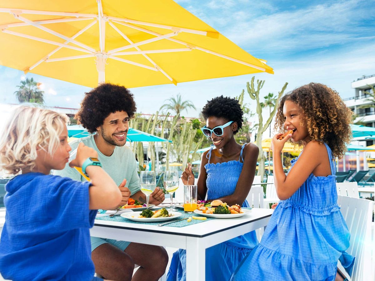 A family of four eating at a white table under a yellow parasol on a poolside terrace, with palms and turquoise parasols behind them.