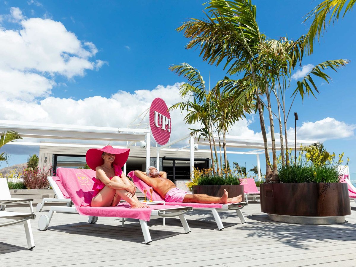 Two adults on pink-towelled sun loungers on a timber roof deck beside potted palms, under a large pink UP! sign and a blue sky.