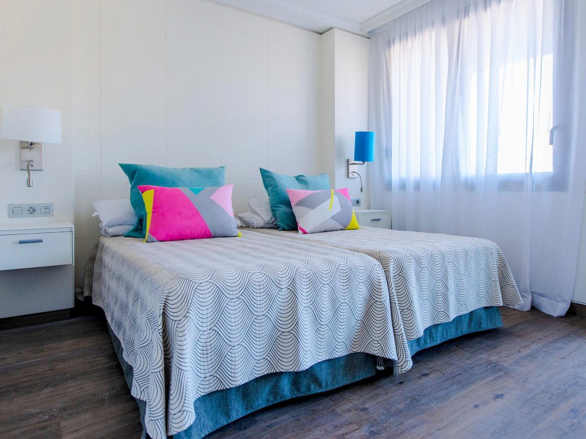Bedroom with two single beds pushed together under a grey cover, pink and turquoise cushions, wall-mounted reading lights and a bright curtained window.