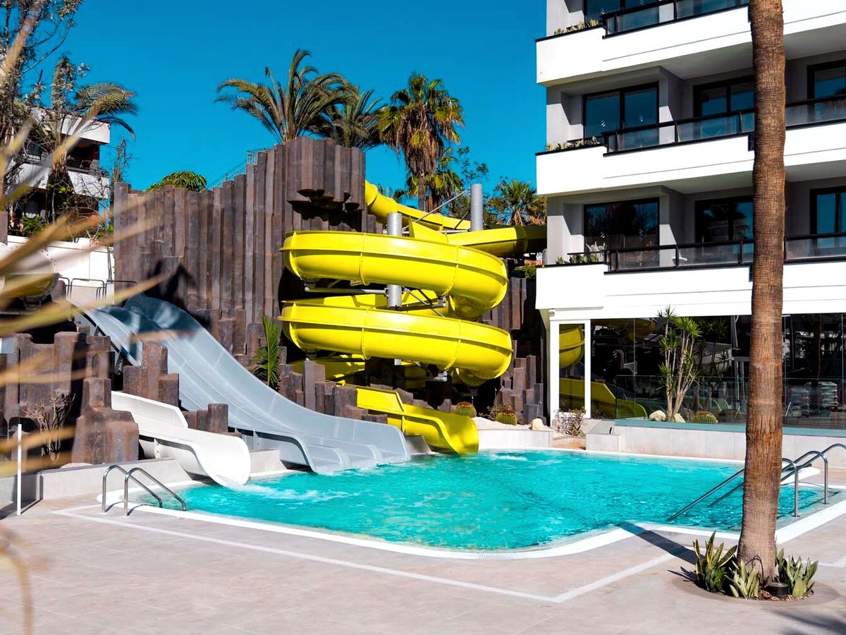 A yellow spiral flume and two open grey slides running down a dark rock-effect wall into their own turquoise pool, with palms and hotel balconies behind.