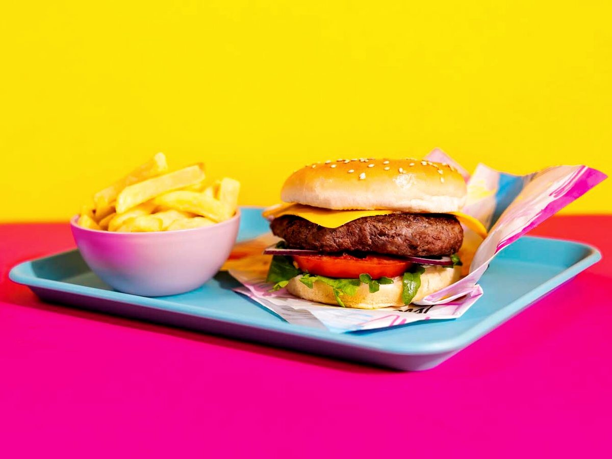 A cheeseburger on a blue tray with a bowl of chips, against a pink and yellow background.