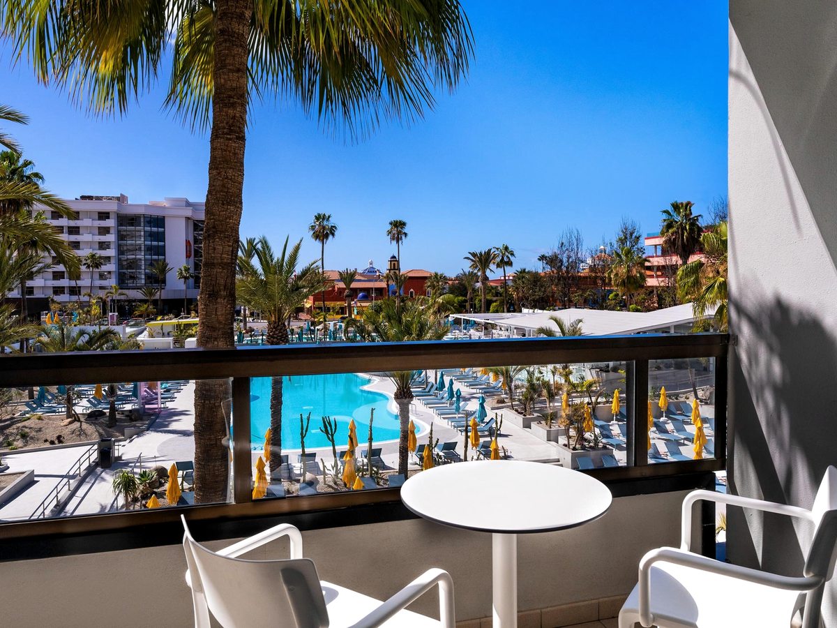 Balcony with a white table and two chairs behind a glass balustrade, looking across palms to the hotel's curved pool and rows of yellow parasols.