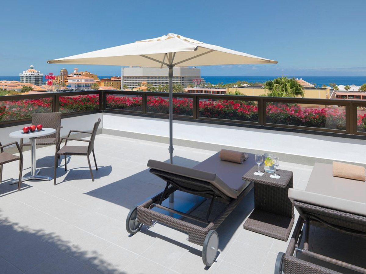 Large private room terrace with wheeled sun loungers, a cream parasol and a bistro table and chairs behind a flower-filled balustrade, with resort rooftops and the sea beyond.
