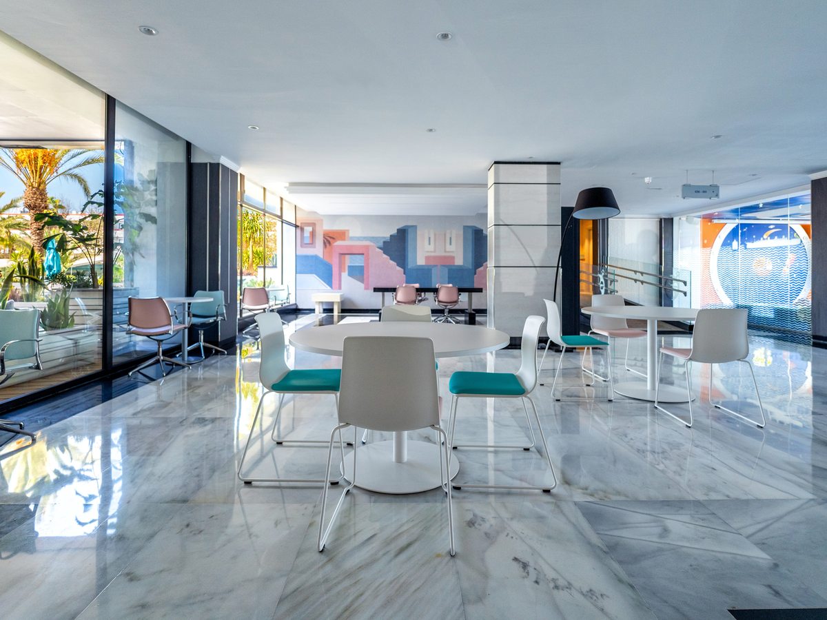 Hotel lobby with round white tables and pastel and teal chairs on a pale marble floor, a colourful geometric mural on the far wall and full-height windows onto the gardens.