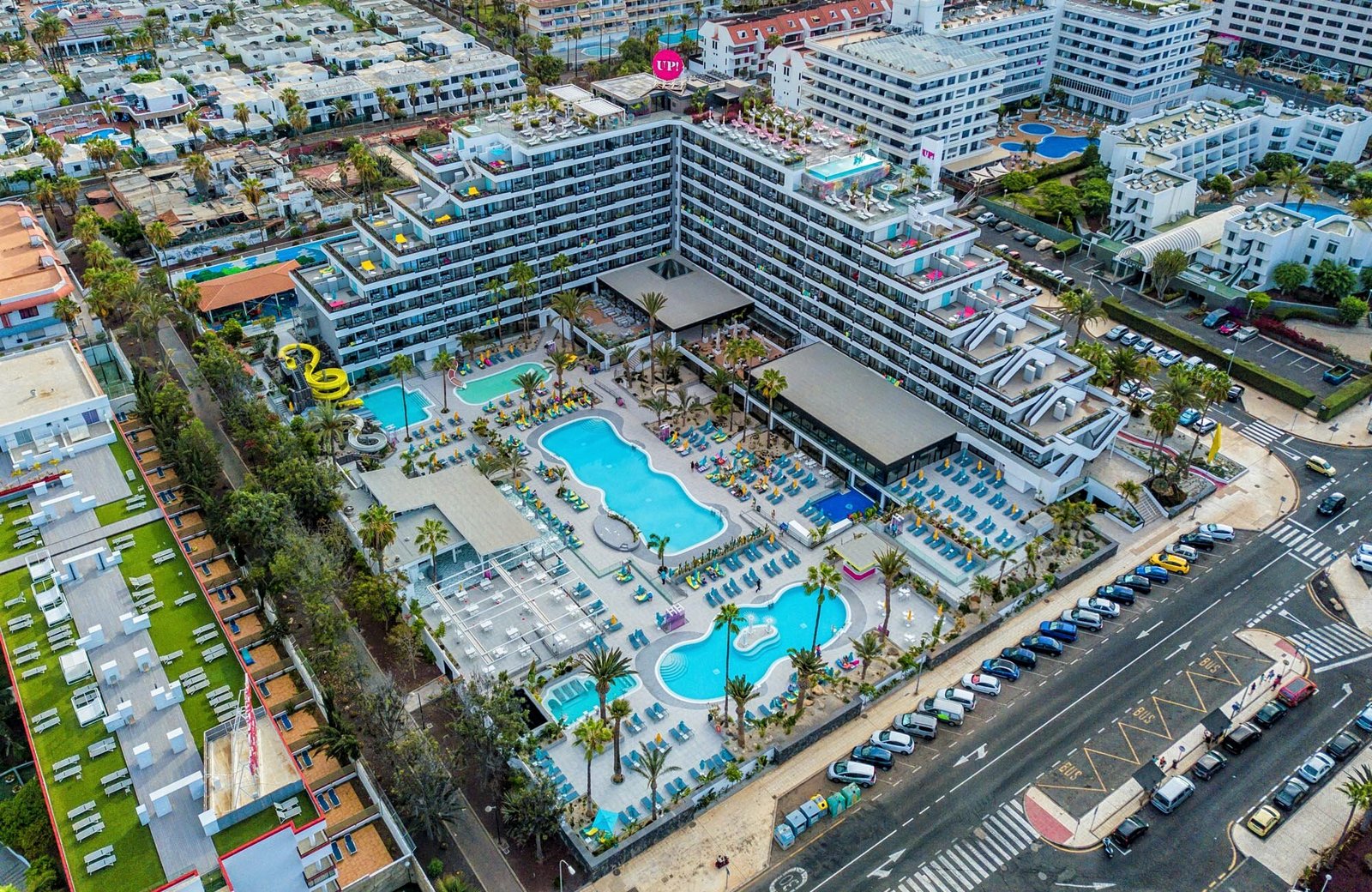 Aerial view of Spring Hotel Bitácora: a white high-rise wrapped around a terrace of pools, water slides and palms, with the streets and low-rise buildings of Playa de las Américas all around it.