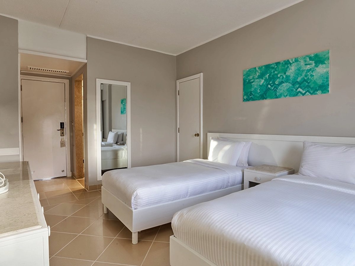 A hotel room with two single beds side by side under a turquoise painting, a white bedside cabinet between them, a white dresser in the foreground and a mirror beside the entrance door.