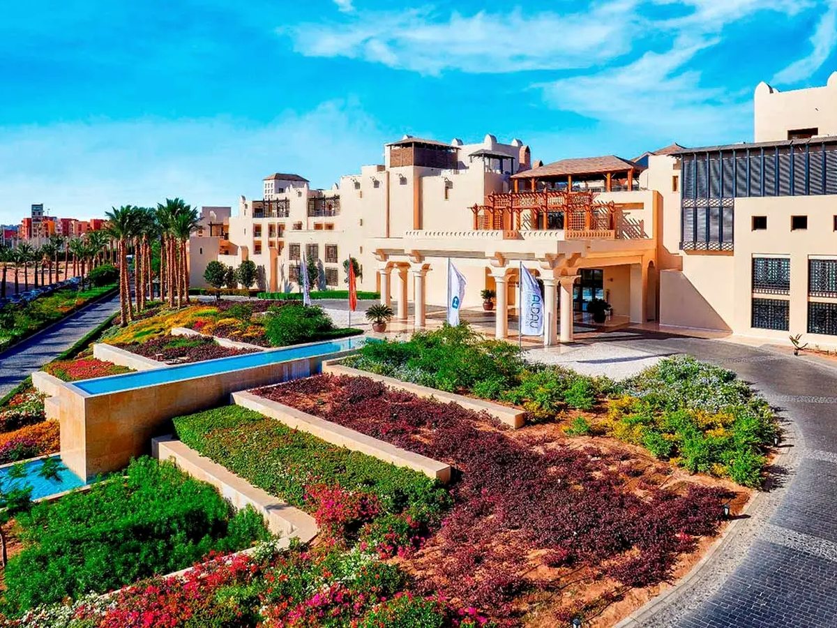 A cream-stone hotel entrance with a columned portico and flagpoles, fronted by terraced beds of red and green planting, narrow water channels and date palms under a blue sky.