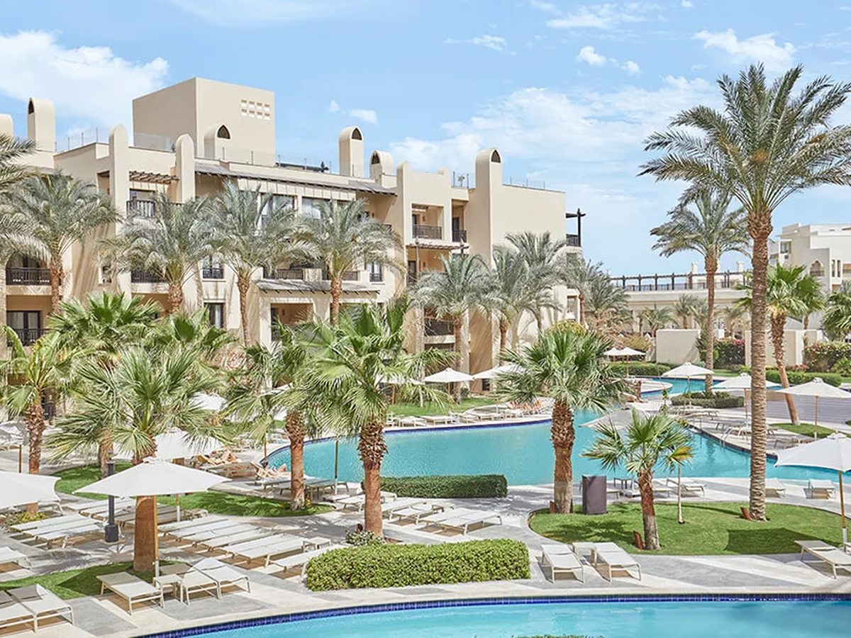Cream low-rise hotel buildings behind a curving blue pool, with palms, lawns, sun loungers and parasols.