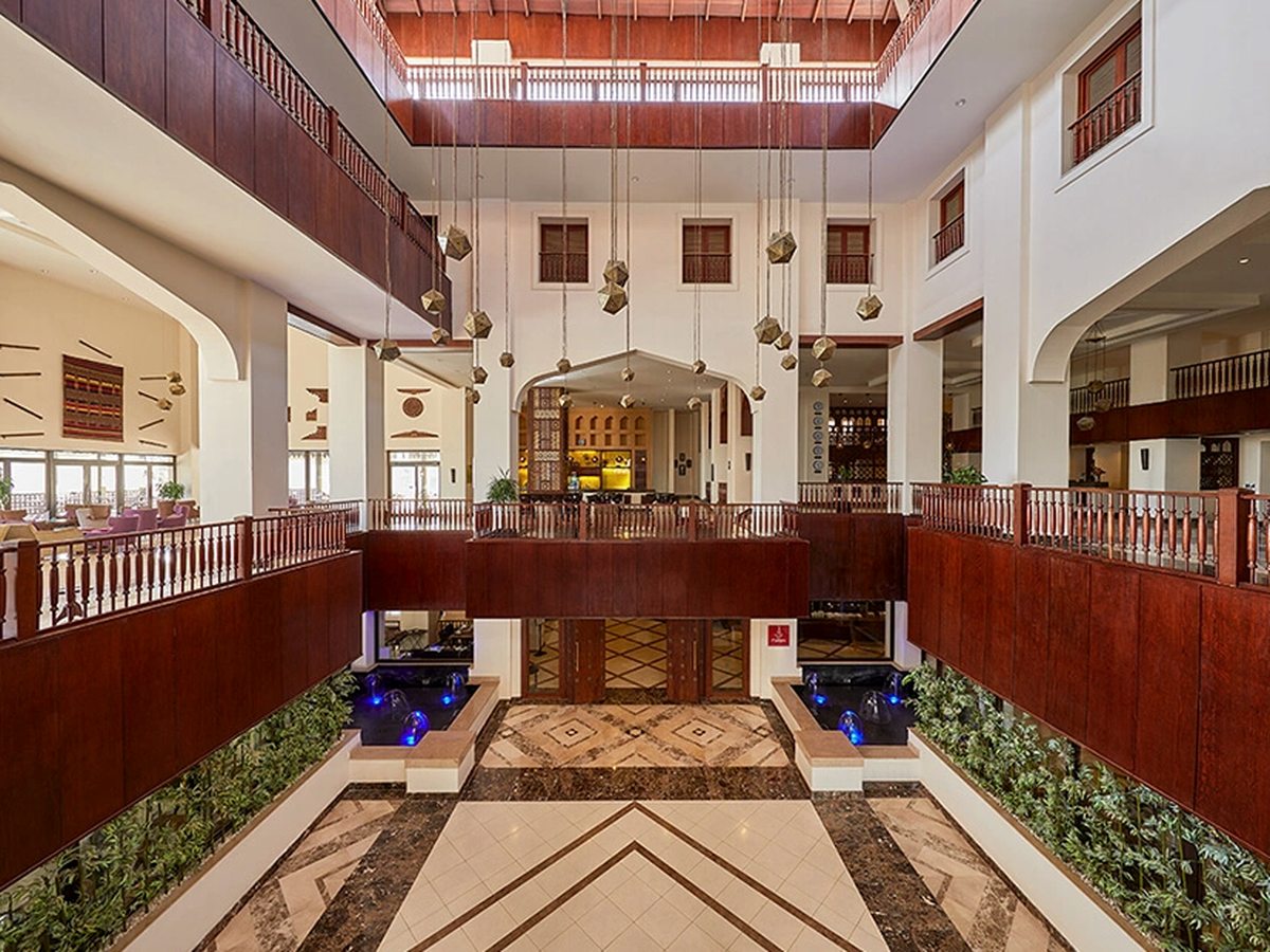 A multi-level atrium lobby with wooden balustrades, arched openings, planted urns and a patterned tiled floor.