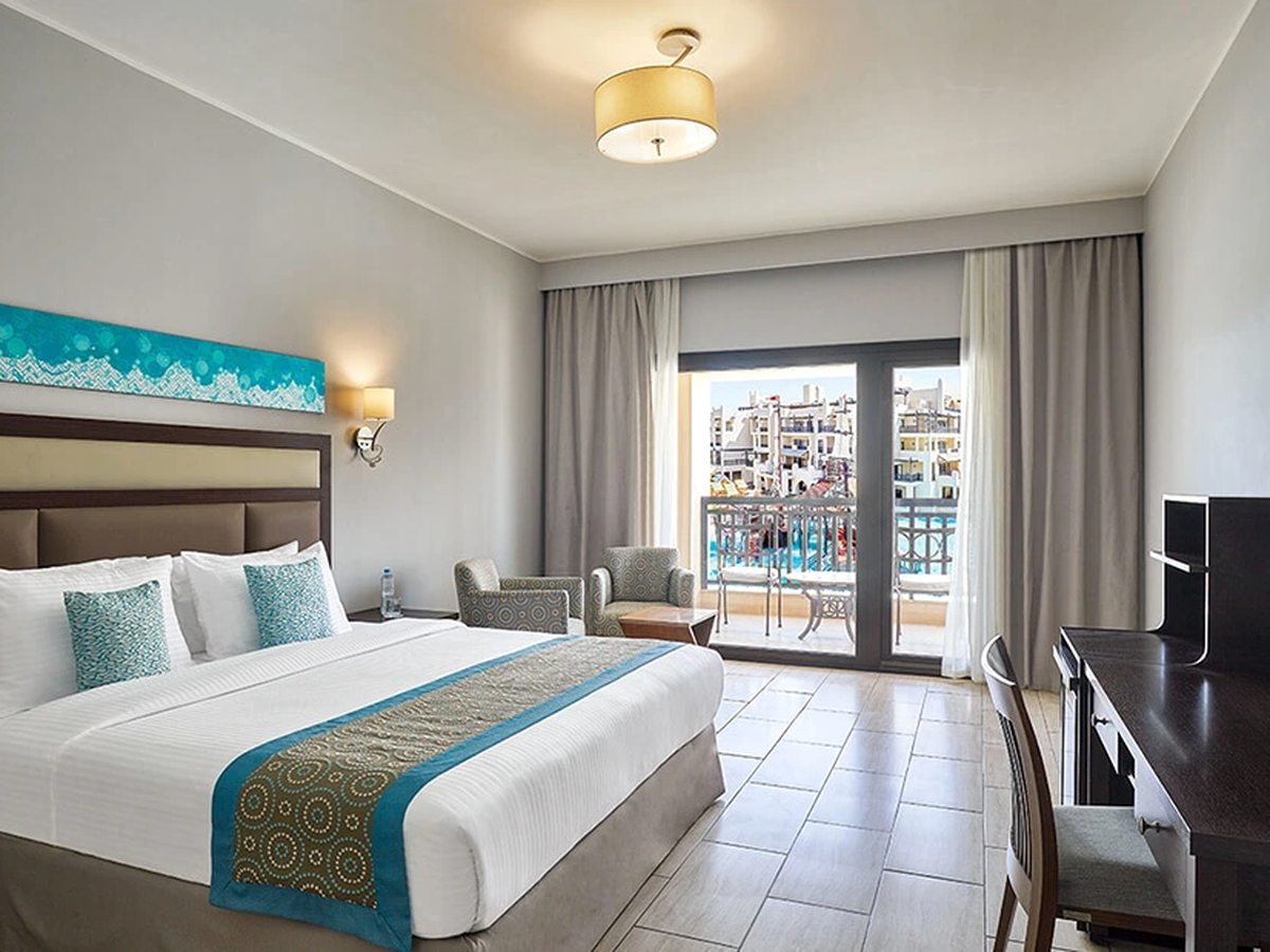 A room with a king bed dressed in white with a turquoise runner, a desk and television, and open doors onto a furnished balcony.
