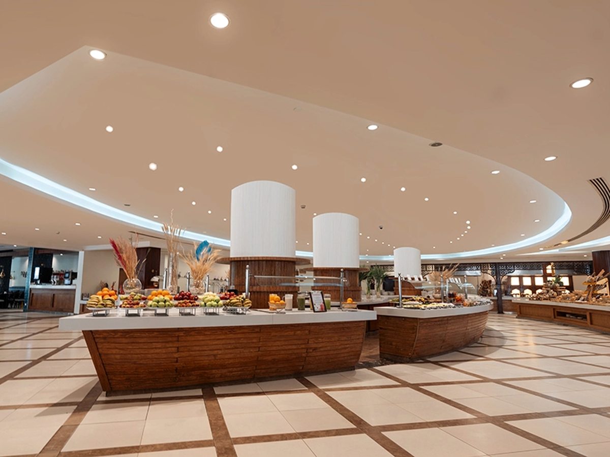 A large buffet hall with curved wooden serving counters holding fruit and pastries, a cream tiled floor and a domed, lit ceiling.