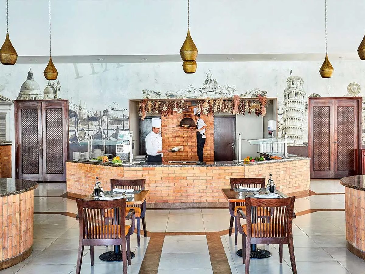 A brick-fronted restaurant counter with chefs working behind it, a mural of Italian landmarks above and set dining tables in front.
