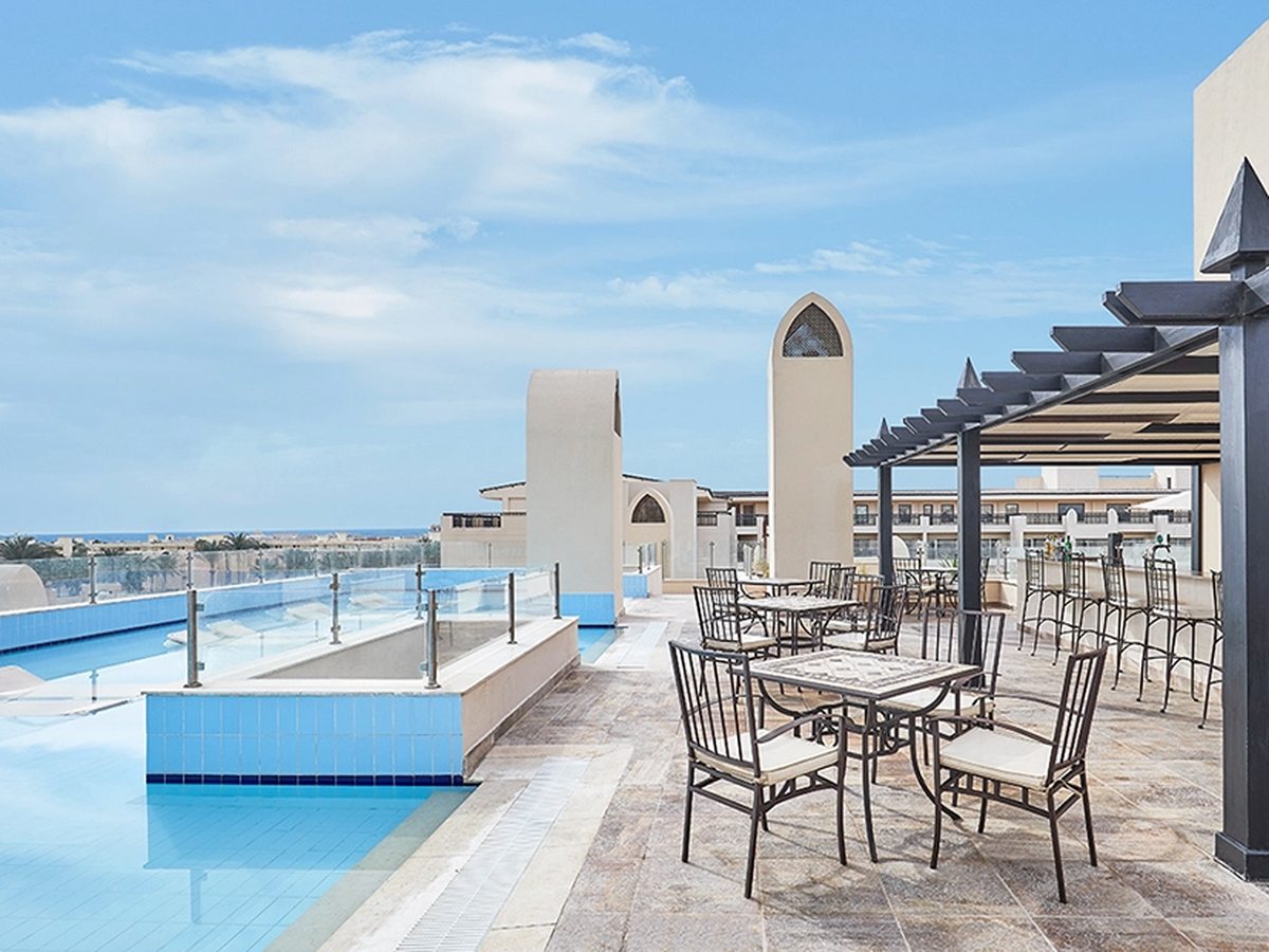 A rooftop pool behind a glass balustrade, with a shaded bar, stools and tables on the terrace and the sea on the horizon.