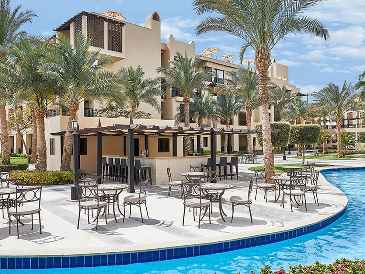 A shaded poolside bar with high stools and metal tables on a terrace beside a curving blue pool, backed by palms and cream hotel buildings.
