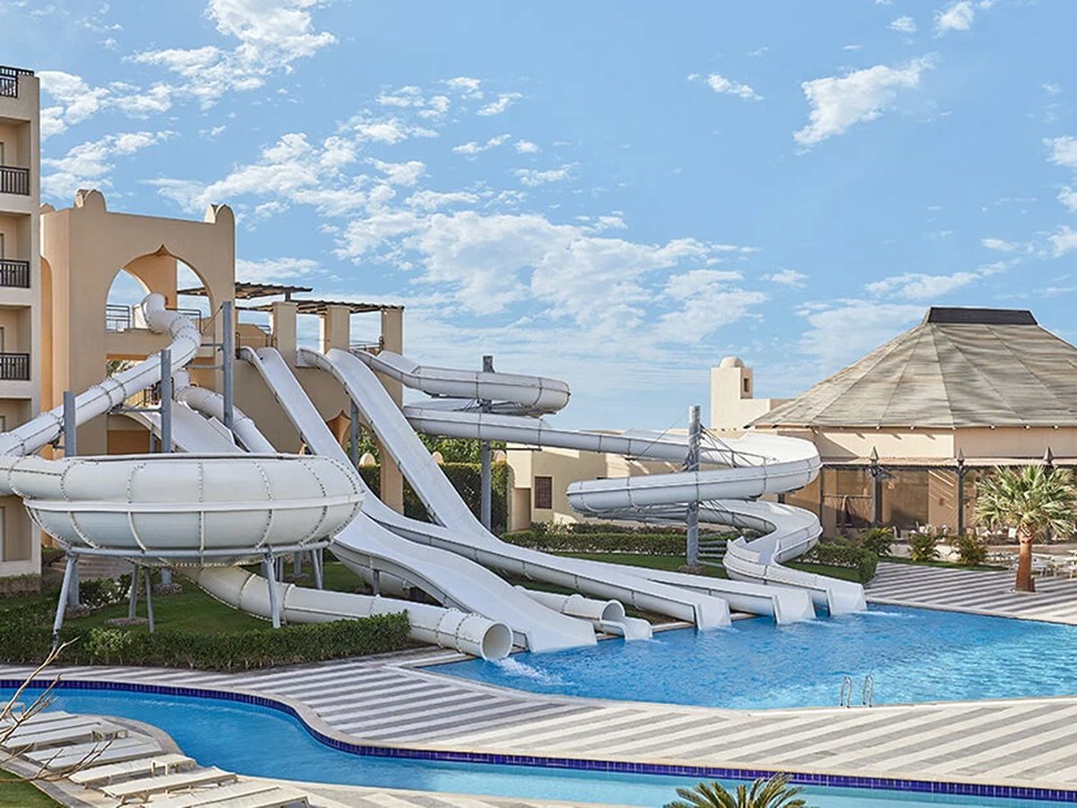 A cluster of white water slides, including a wide bowl and a spiral tube, running down into a blue pool beside cream hotel buildings.
