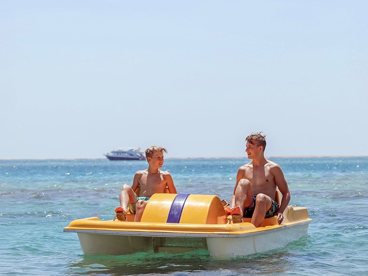 Two people on a yellow pedalo on flat blue sea, with a small boat in the distance.