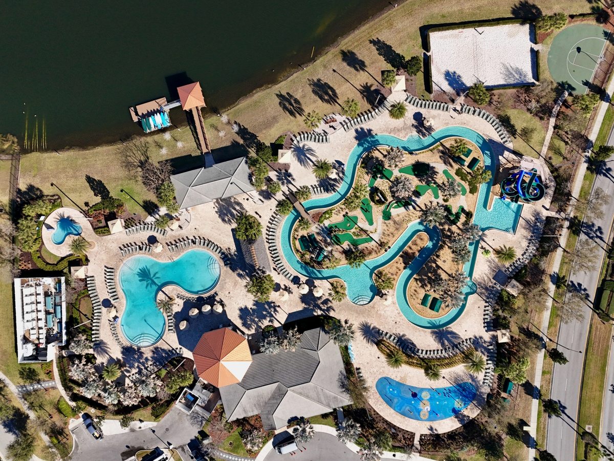 Overhead aerial of a water park with a winding lazy river, a rounded pool, curved waterslides and a splash pad, beside a lake.