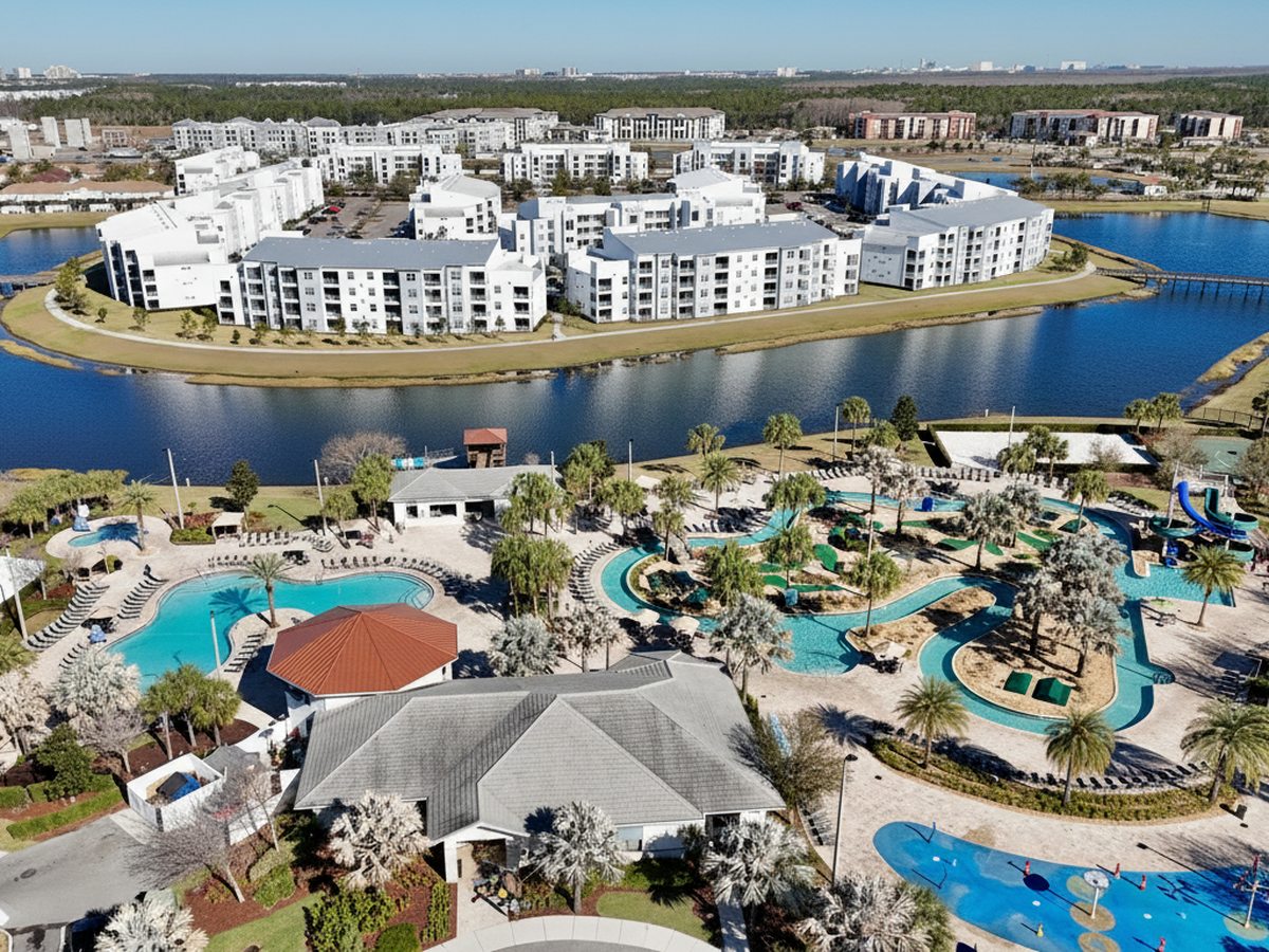 Aerial view of a resort with a large blue water park in the foreground, a splash pad, lazy river and waterslides, backed by rows of white apartment buildings and a lake.