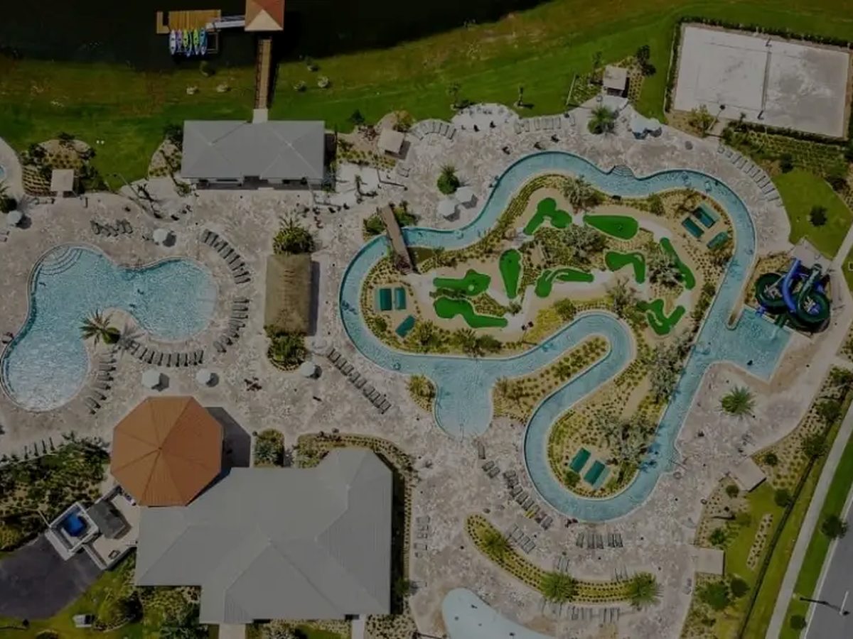 Top-down aerial of a water park with a freeform pool, lazy river, blue waterslides and a sports court beside a clubhouse and a road.