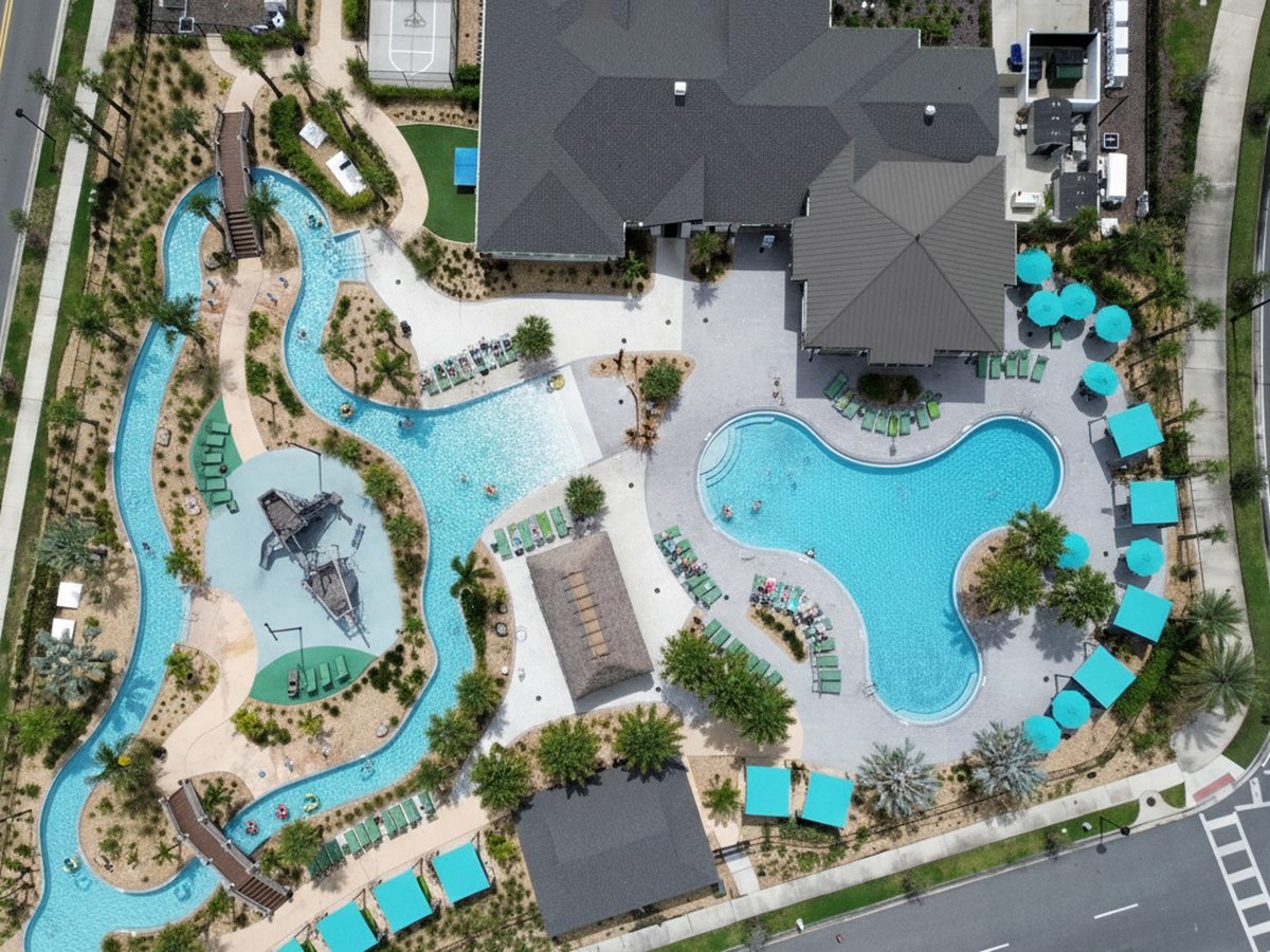 Overhead aerial of a freeform pool and lazy river with turquoise parasols, poolside cabanas and a sun deck beside a clubhouse.