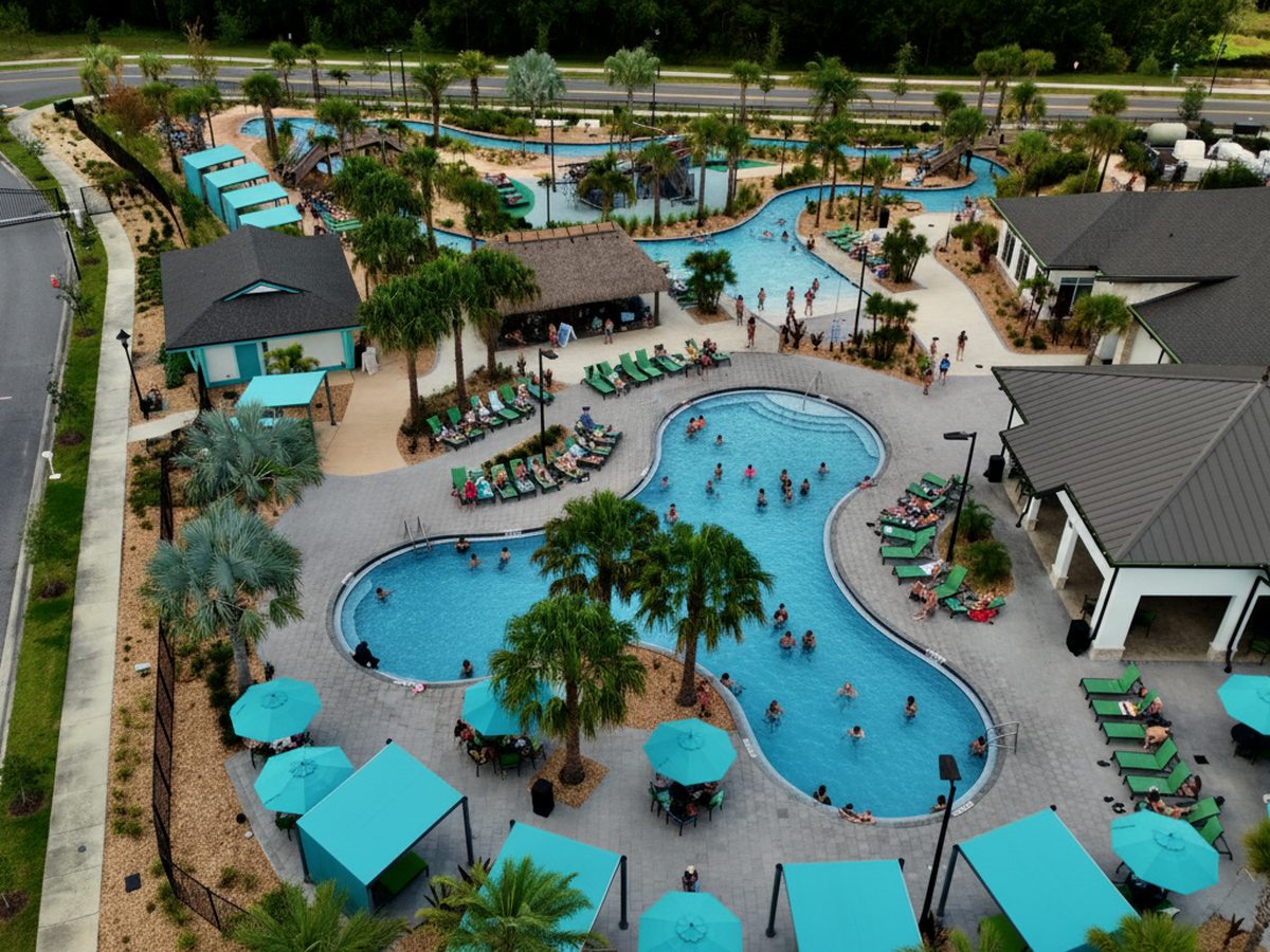 A freeform blue pool with a planted island, green sun loungers and turquoise parasols, with people swimming and a clubhouse behind.