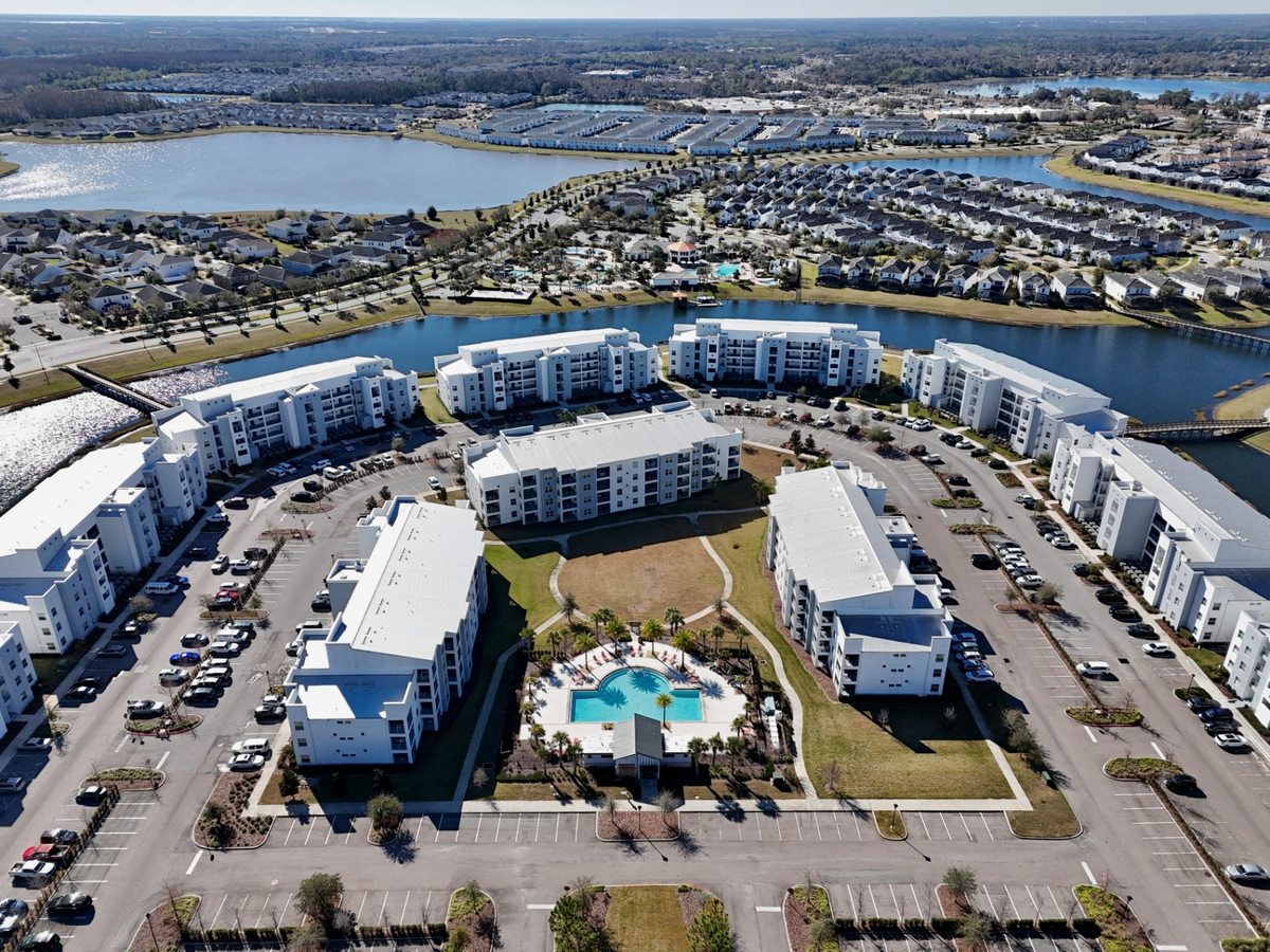 Wide aerial of a resort of multi-storey white apartment blocks around roads and car parks, with a central pool and a canal and lake to one side.