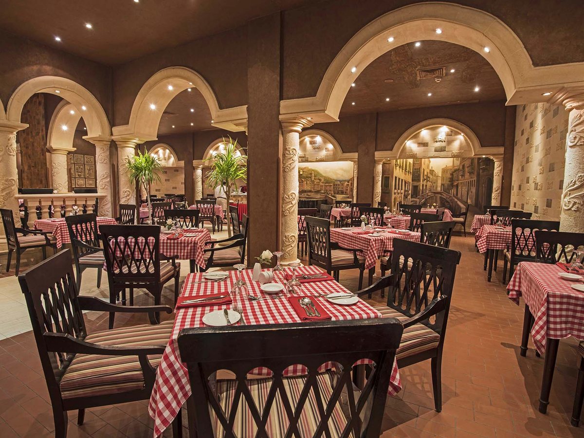 A restaurant with red and white checked tablecloths, cream stone arches, painted Venetian canal scenes and dark wooden chairs.