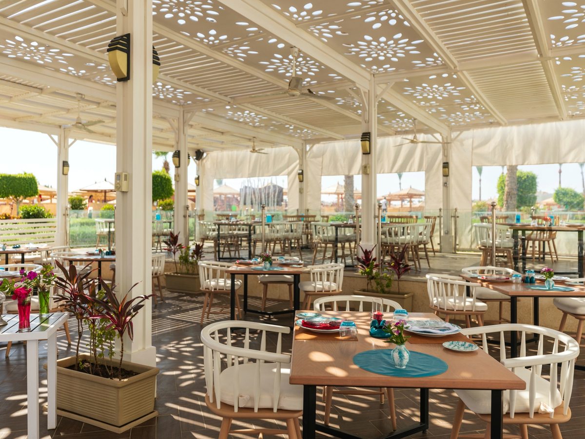 An open-sided restaurant terrace under a white fretwork canopy, with pale wooden tables, round-backed white chairs and gardens beyond.