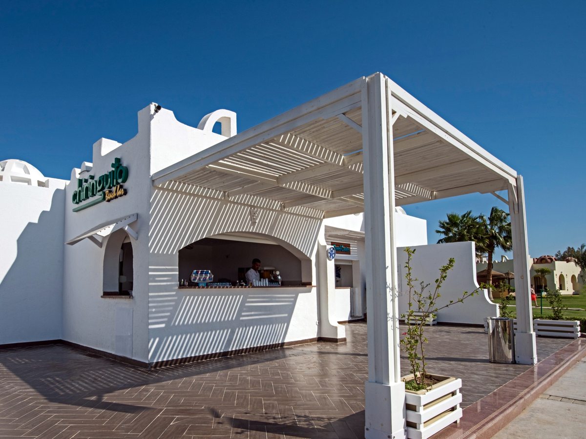 A whitewashed beach bar with a green Chiringuito sign, a slatted white pergola over a tiled terrace and a barman behind the counter.