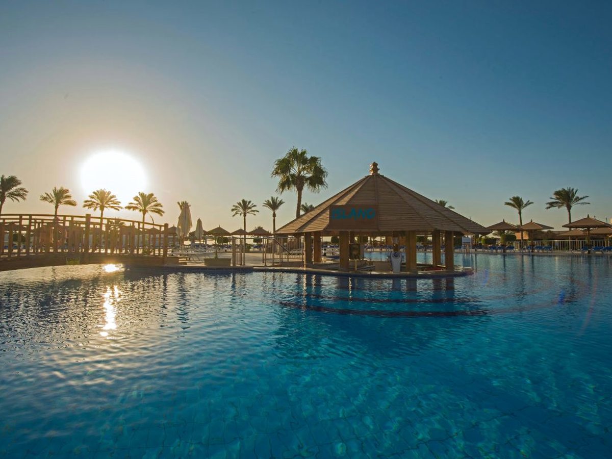 A thatched-roof pool bar signed Island standing on a platform in the middle of a large pool, with palms and a low sun behind.