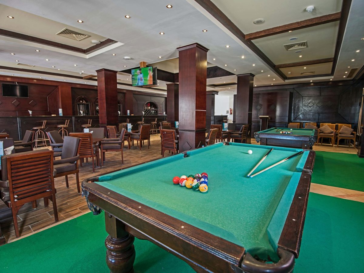 A pub interior with a full-size billiards table on green carpet, a dark wood bar counter, wooden chairs and a wall-mounted television.