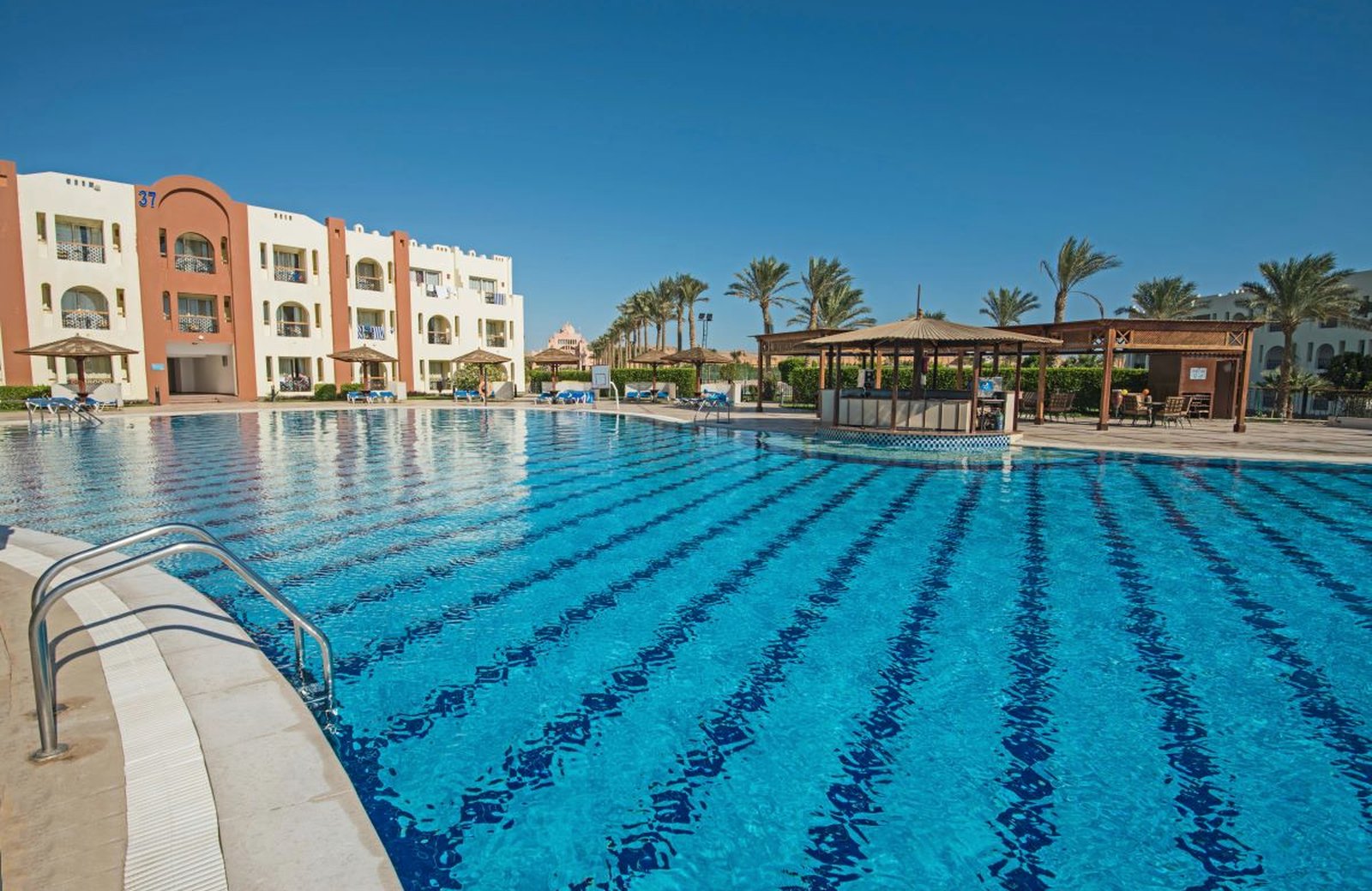A large blue pool at SUNRISE Royal Makadi with a thatched pool bar to one side and white and terracotta guest blocks behind, under a clear sky.