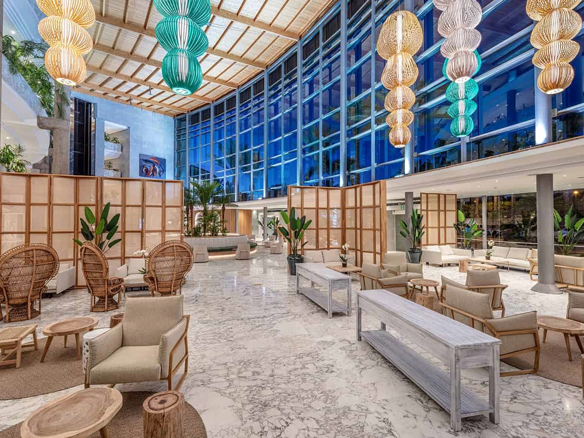 Tall, light lobby lounge with pale linen armchairs, wicker peacock chairs, wooden screen dividers and clustered lantern pendants above a marble floor, with a full-height glass wall behind.