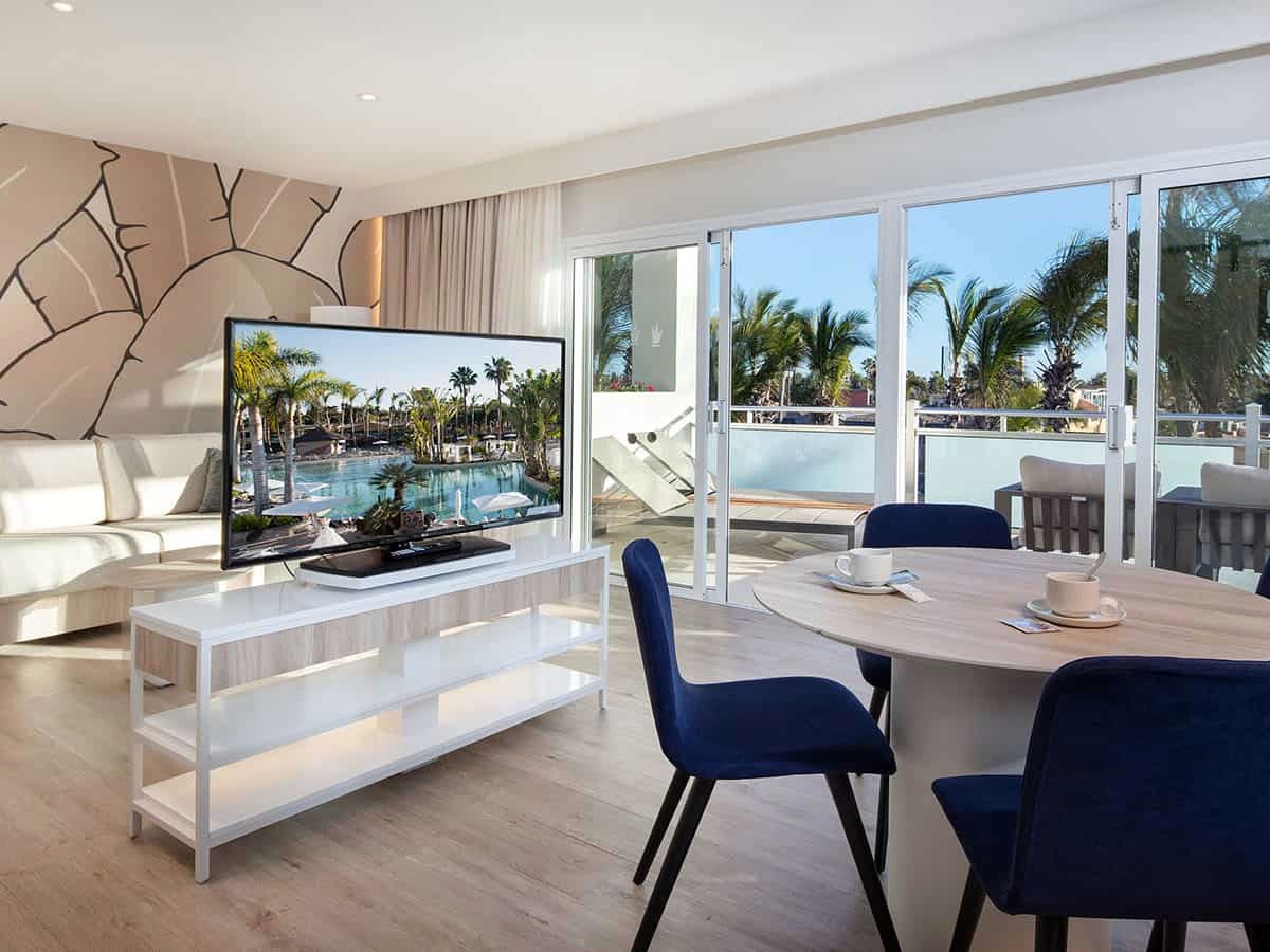 Suite living area with a white corner sofa, a television on a low white unit, a round dining table with navy velvet chairs and sliding doors onto a furnished balcony above the palm gardens.