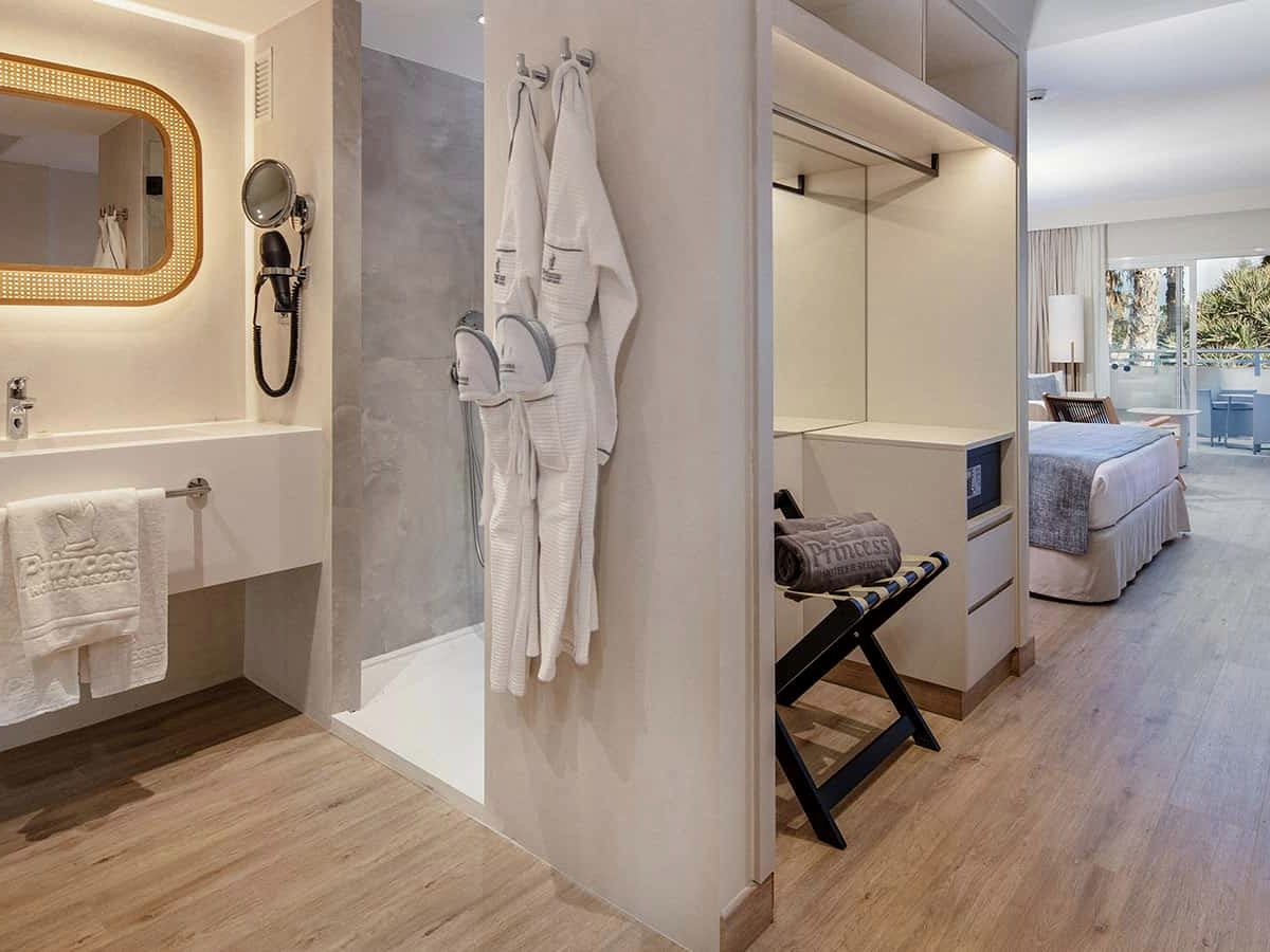 Room bathroom with a long white vanity, a cane-framed lit mirror and an open walk-in shower with bathrobes hung alongside, opening past a wardrobe and luggage rack into the bedroom.