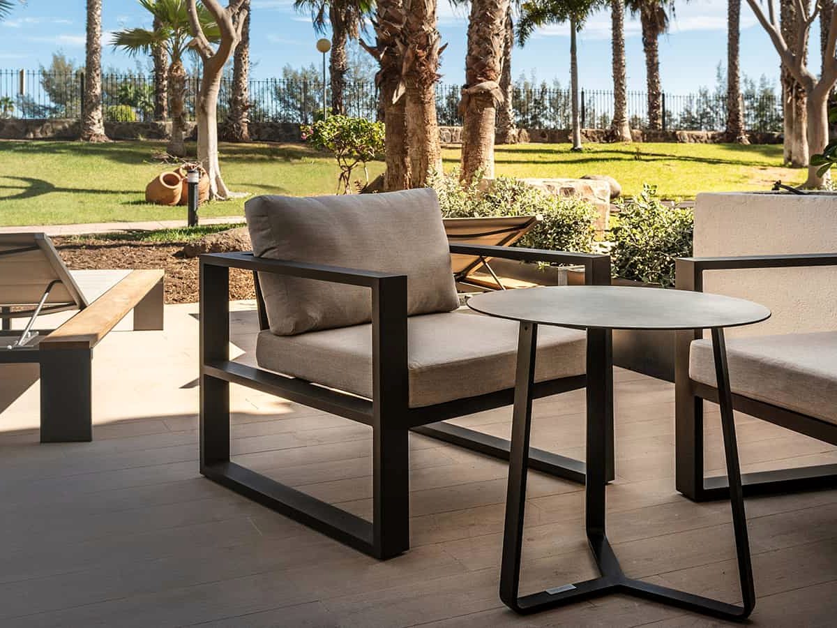 Private ground-floor terrace with grey cushioned armchairs, a round side table and wooden sun loungers, opening onto a lawn planted with palms.