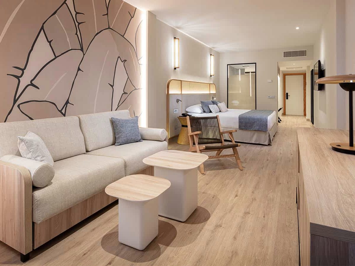 Open-plan hotel room with a cream sofa bed and two low tables in the foreground, a large leaf mural on the wall and a double bed with a rattan headboard and a glass-screened bathroom behind.