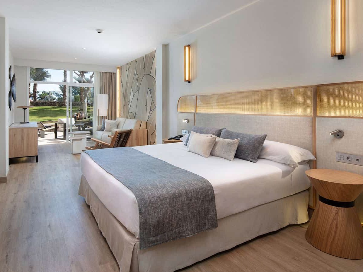 Ground-floor double bedroom with a wide bed, a lit headboard panel and sliding glass doors opening onto a furnished terrace with lawn and palms beyond.