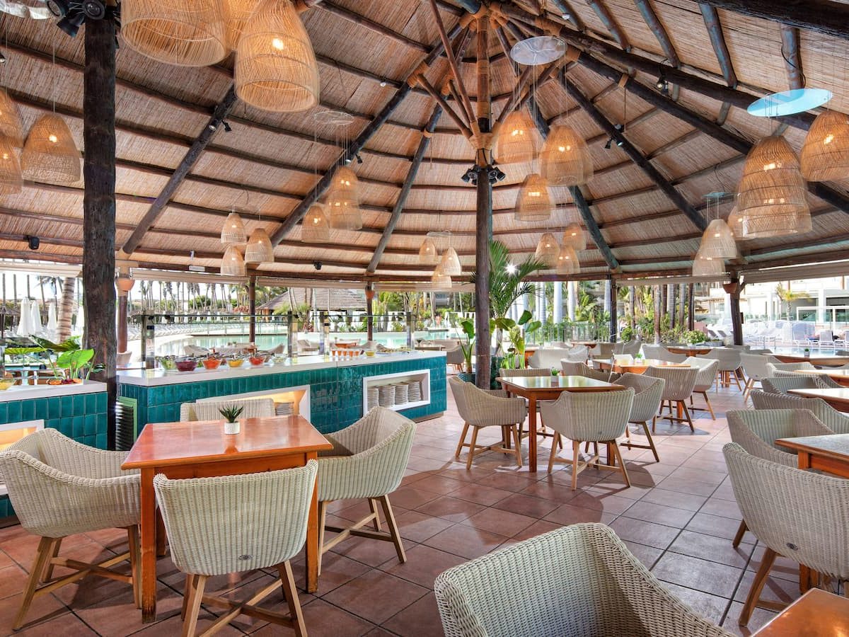 Large thatched-roof poolside restaurant with a turquoise-tiled buffet counter, cream wicker armchairs at wooden tables and woven pendant lampshades, with the pool and palms beyond.