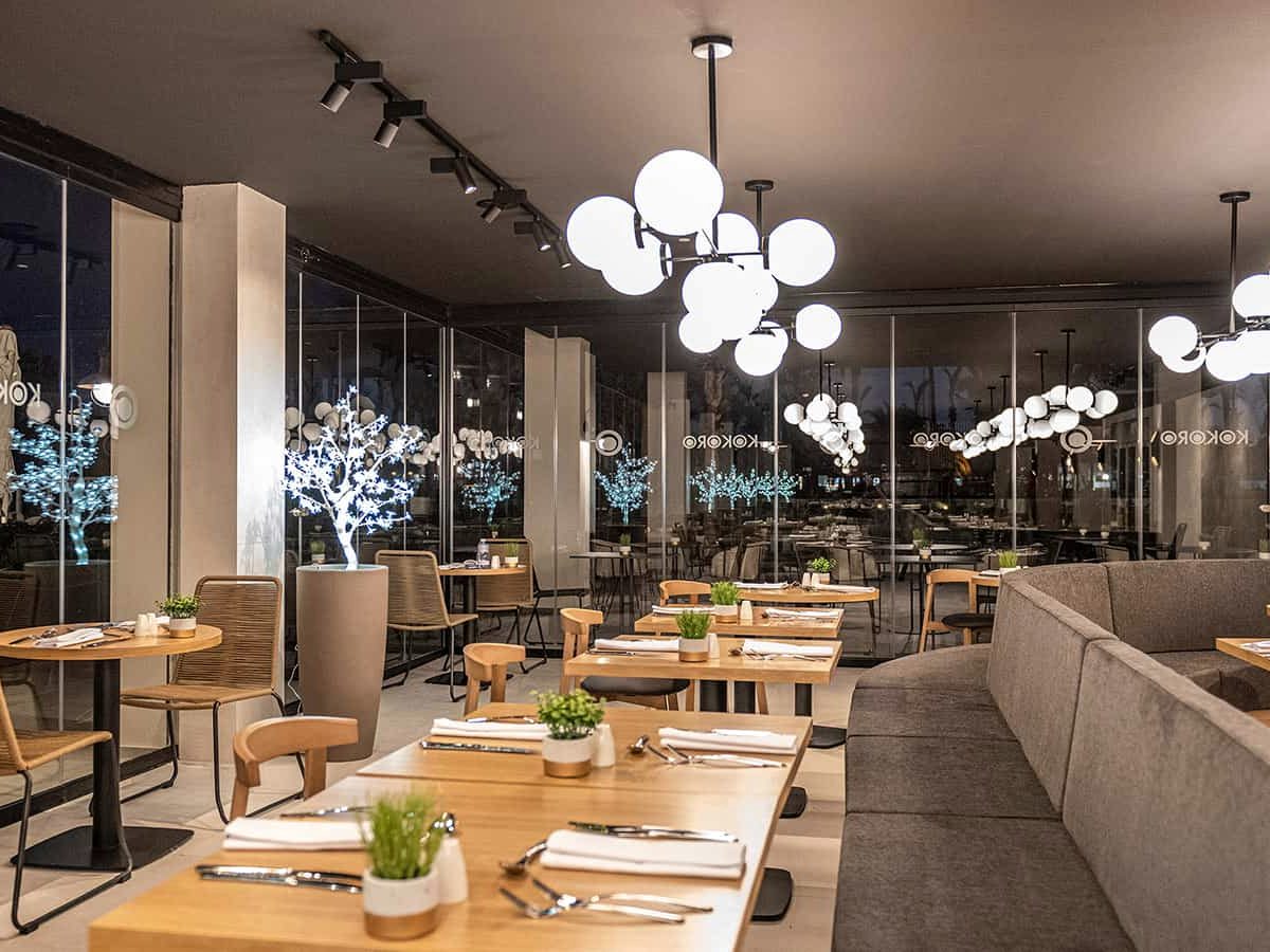 Interior of the Kokoro restaurant with laid wooden tables, cane chairs, a long grey banquette and cluster globe pendant lights, its name etched across the glass wall.