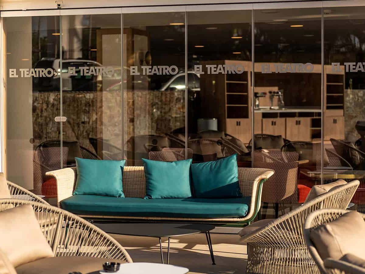Outdoor terrace in front of the El Teatro bar, with cream rope armchairs, a wicker sofa with teal cushions and a low black table, the bar's name lettered across the glass frontage.