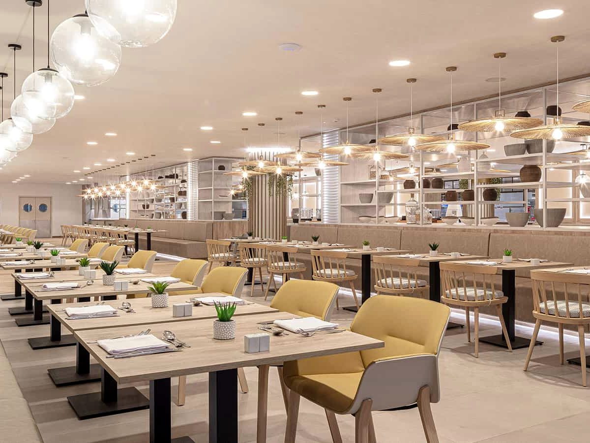 Long, bright restaurant dining room with pale wood tables, yellow and cane chairs, banquette seating and rattan and glass pendant lights, with a service counter at the far end.