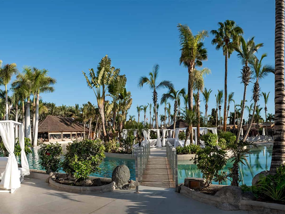 Wooden footbridge with glass balustrades crossing a turquoise pool between planted islands, leading to a paved terrace of white curtained four-poster daybeds under palms.