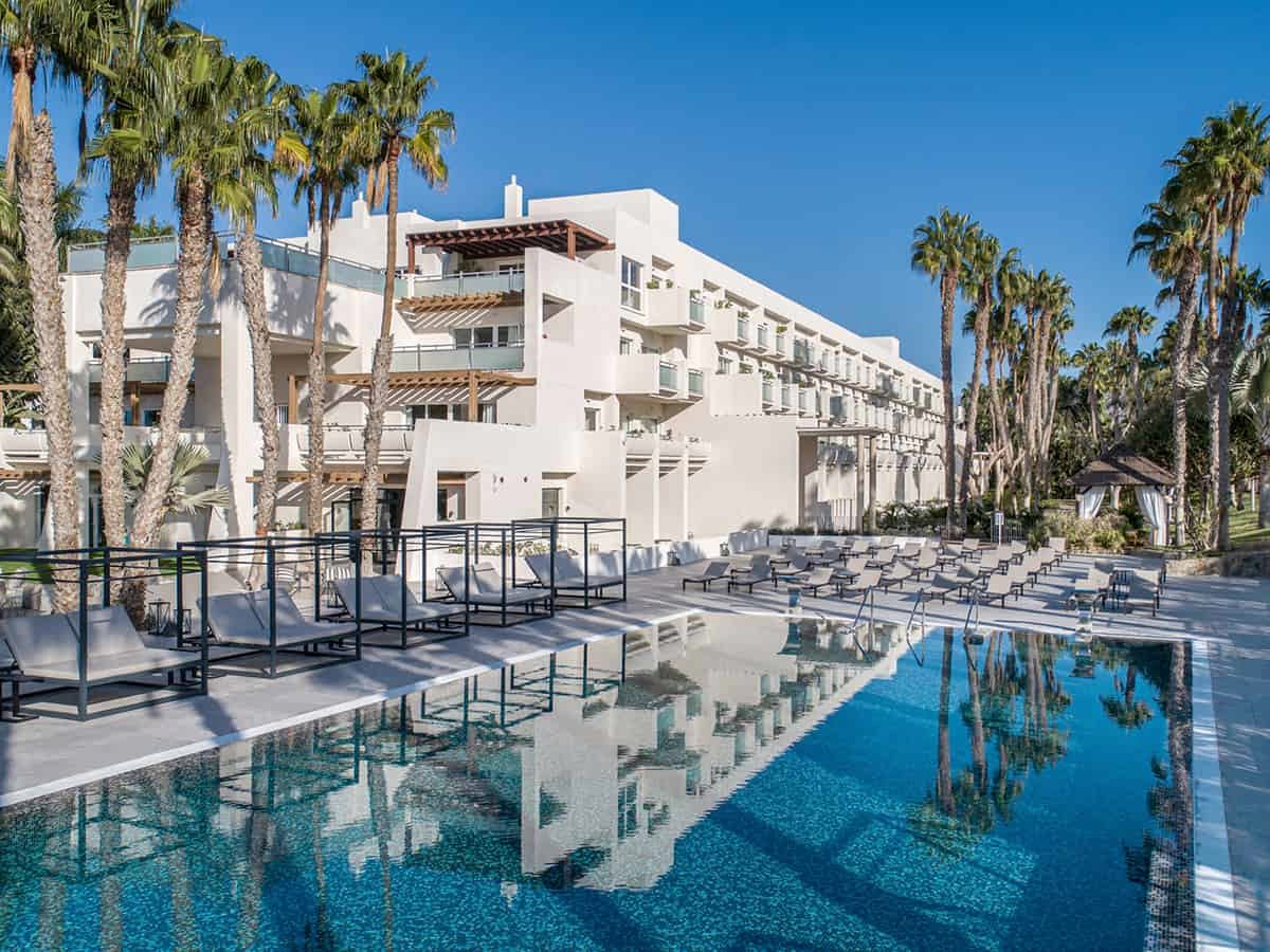 Low white hotel block with balconies and timber pergolas behind a rectangular pool edged with grey sun loungers and tall palms.